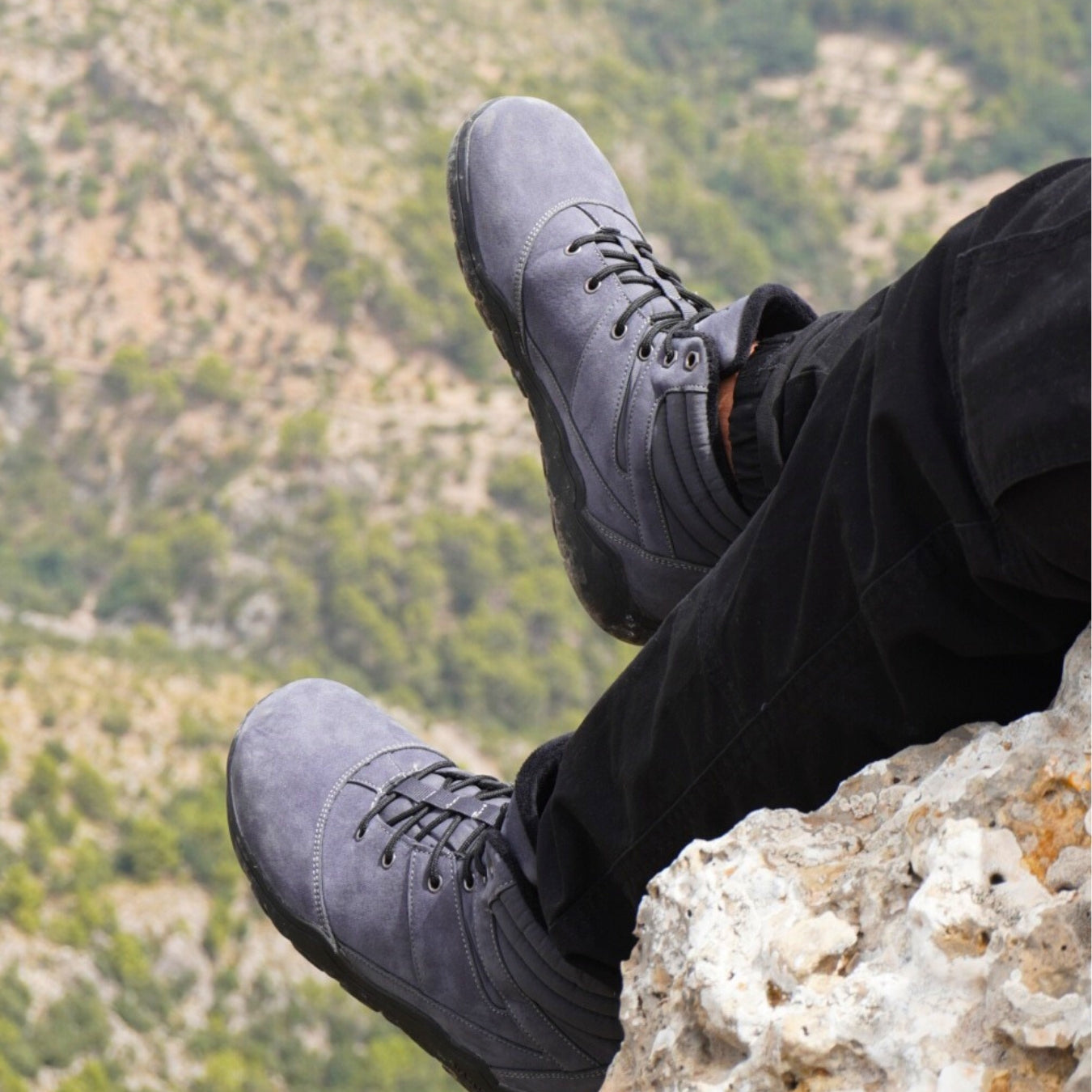 Person wearing gray Naturcontact barefoot hiking boots sitting on a rock ledge with a mountain view, combining grip and natural comfort.