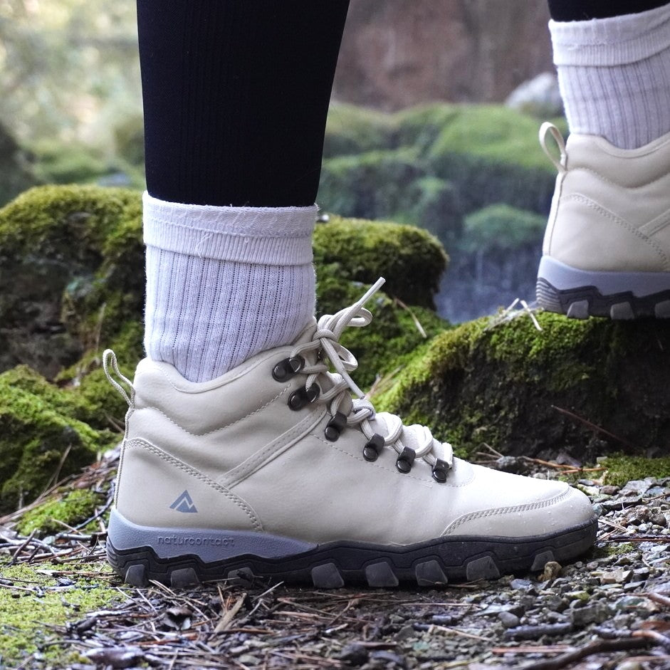 White hiking boots with black soles on a person standing on a mossy ground.
