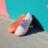 White barefoot shoes with orange soles and purple laces against a colorful wall.