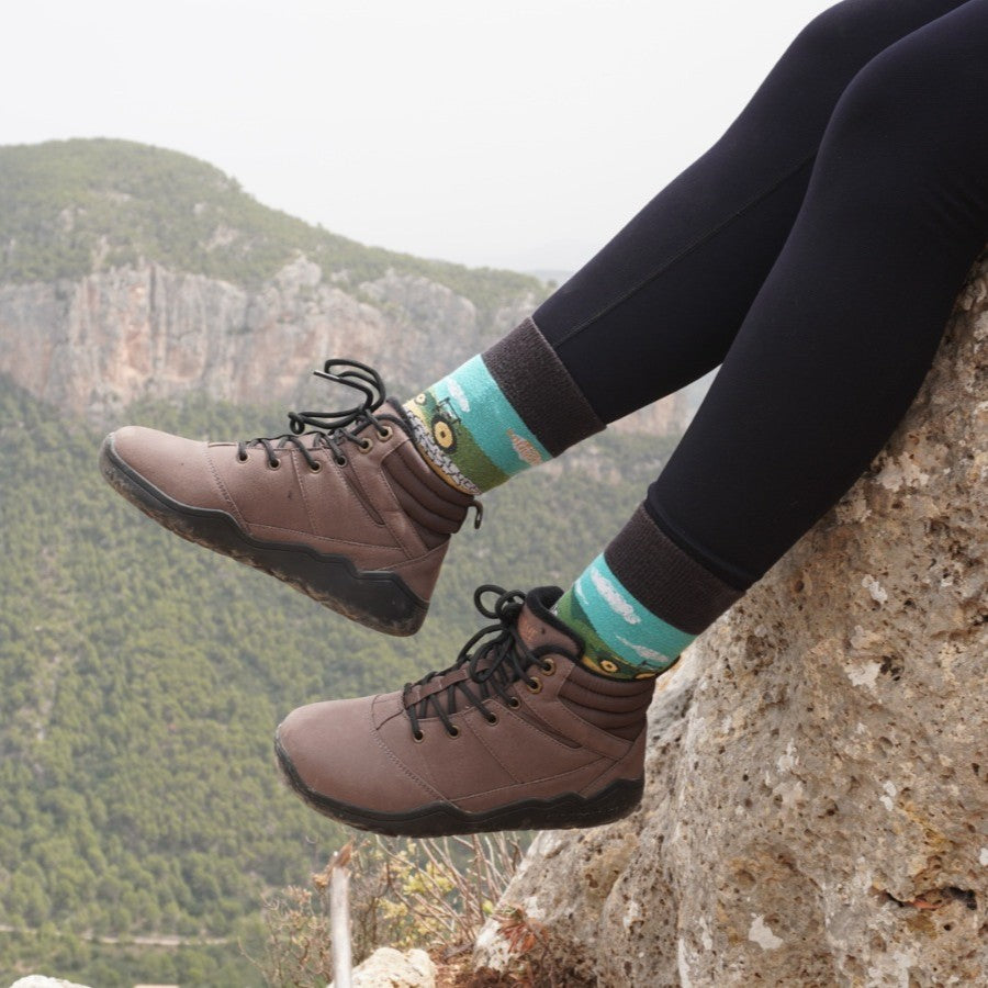 Brown hiking barefoot boots with black soles on a person sitting on a rock with a scenic background.