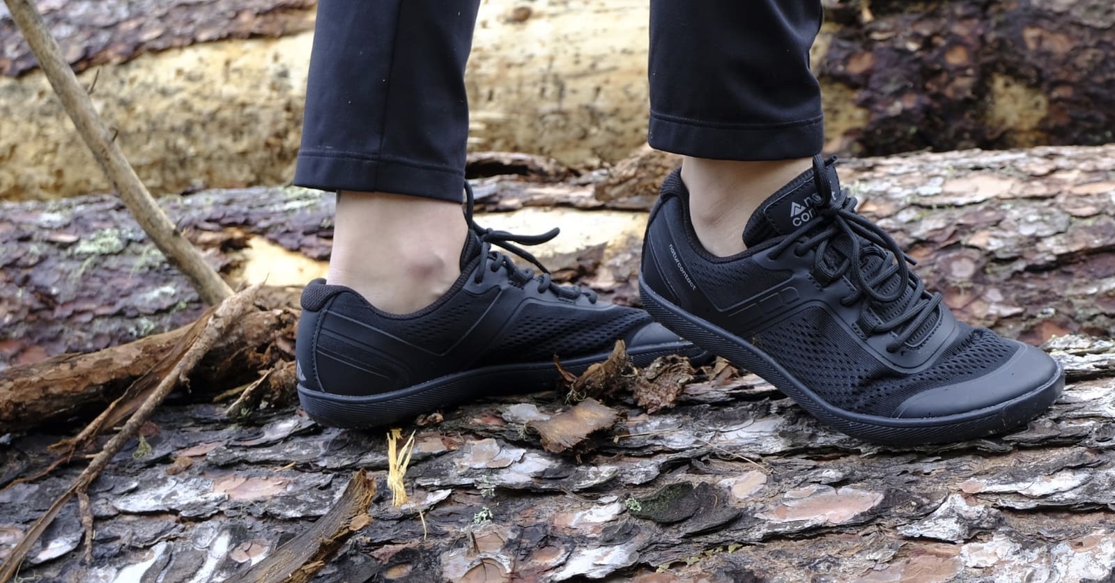 Black barefoot shoes trainers worn on a person standing on a forest floor with logs and leaves.