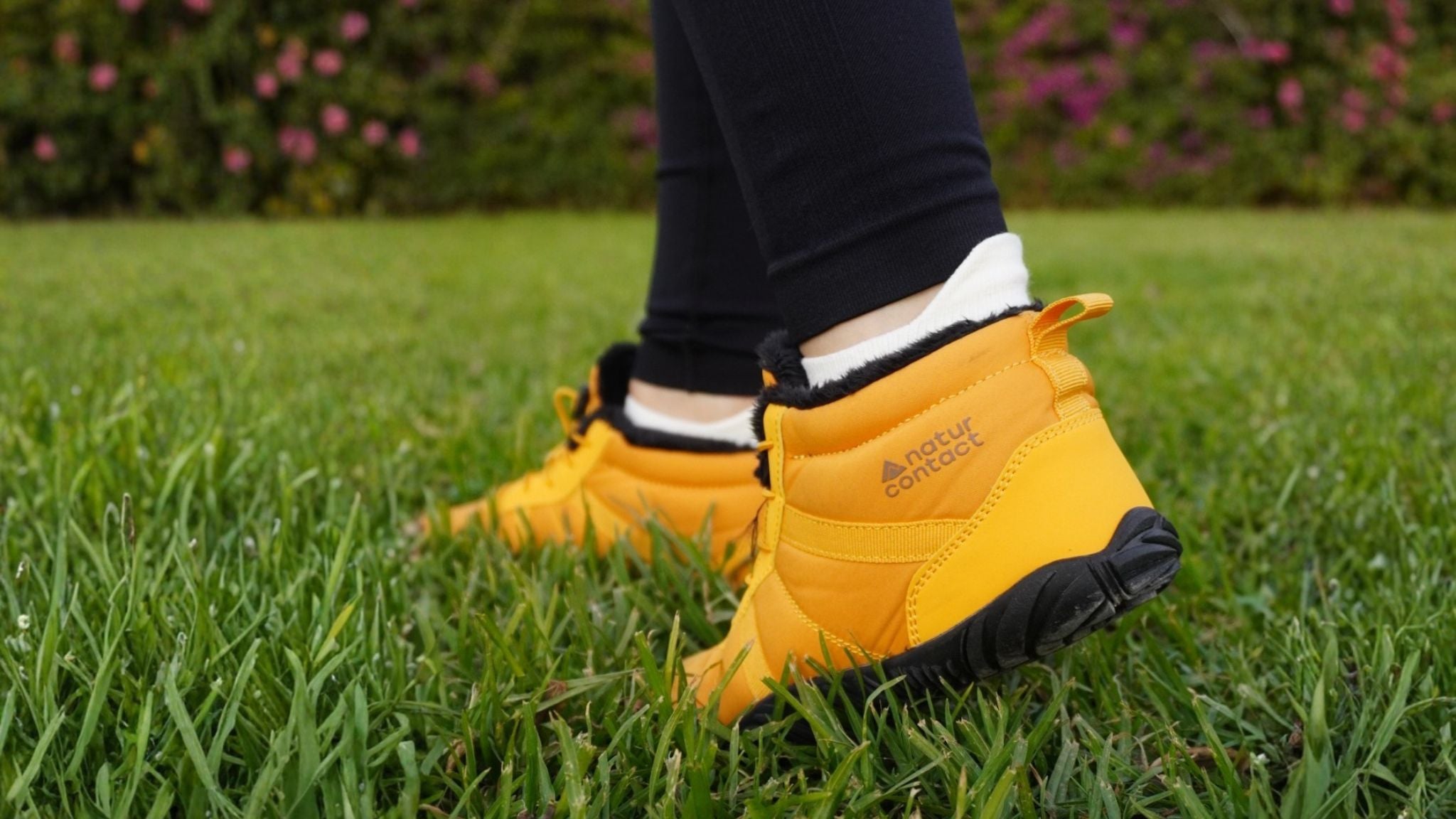 Close-up of a person wearing yellow Naturcontact barefoot shoes standing on green grass, showing the flexible minimalist sole and outdoor-friendly design.