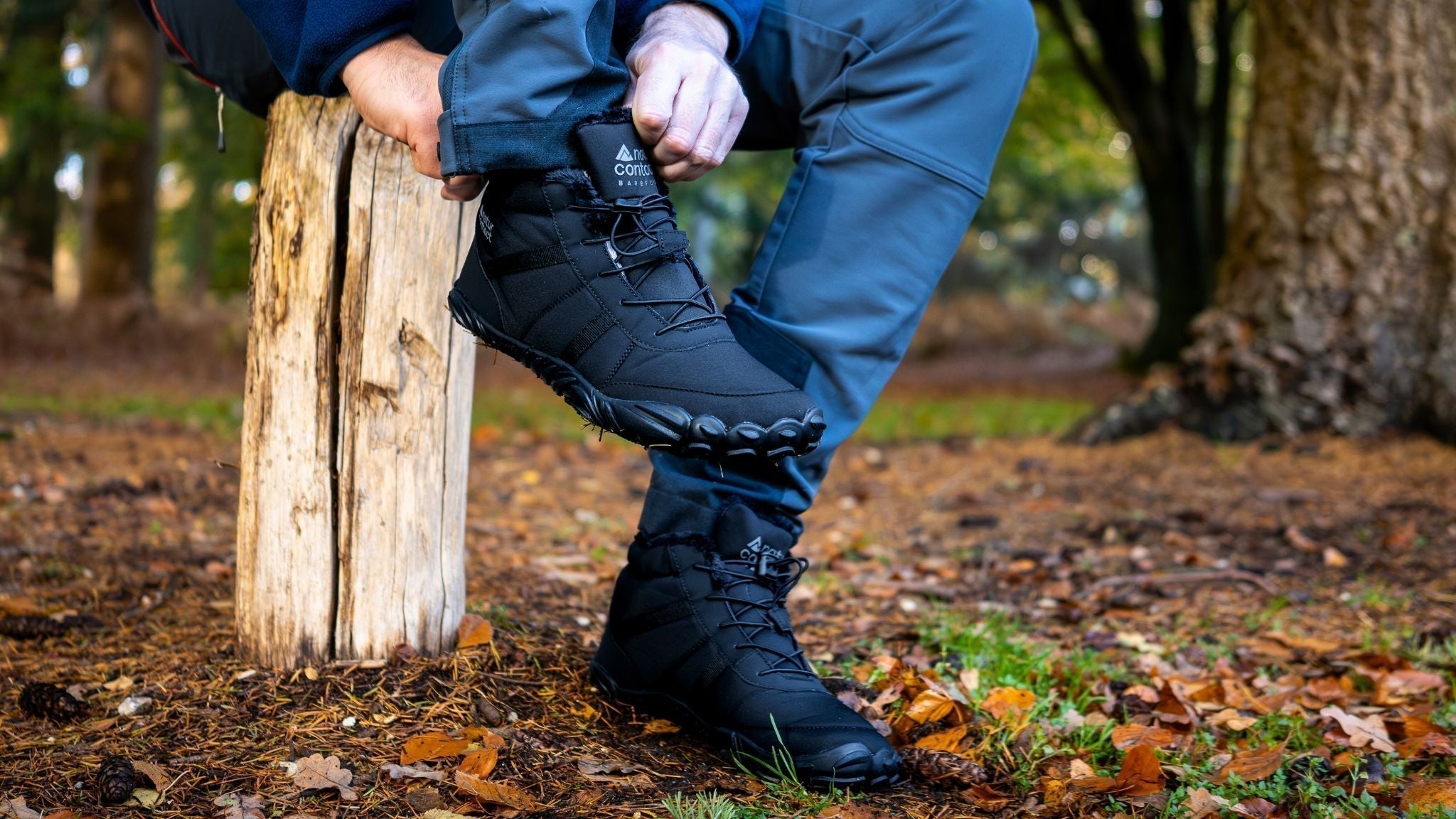 Person sitting on a wooden stump in a forest, adjusting black winter barefoot boots with a flexible sole, designed for natural movement and outdoor comfort.
