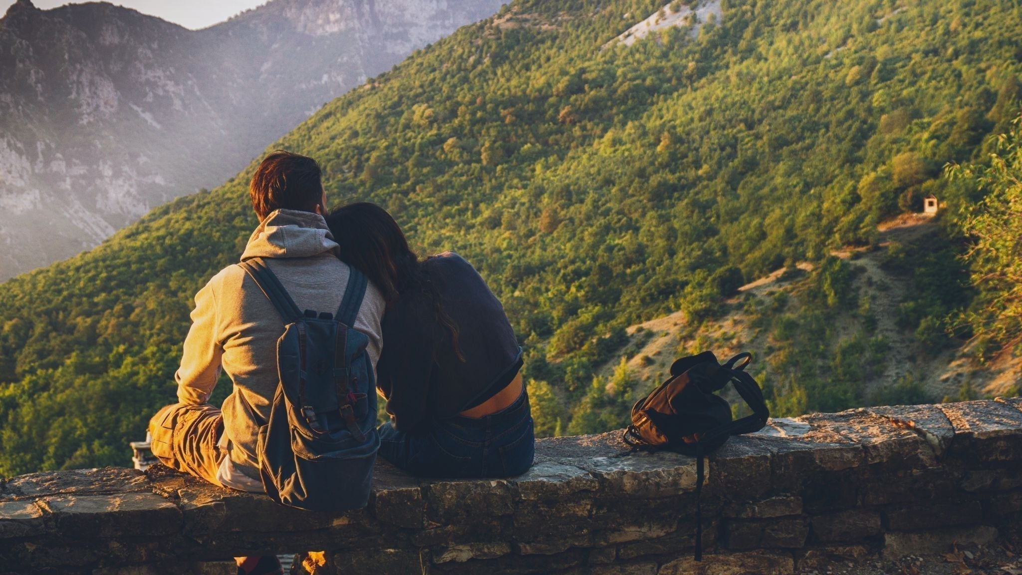 Two people sitting together on a stone wall overlooking a valley and forested mountains during sunset, enjoying a peaceful outdoor moment.