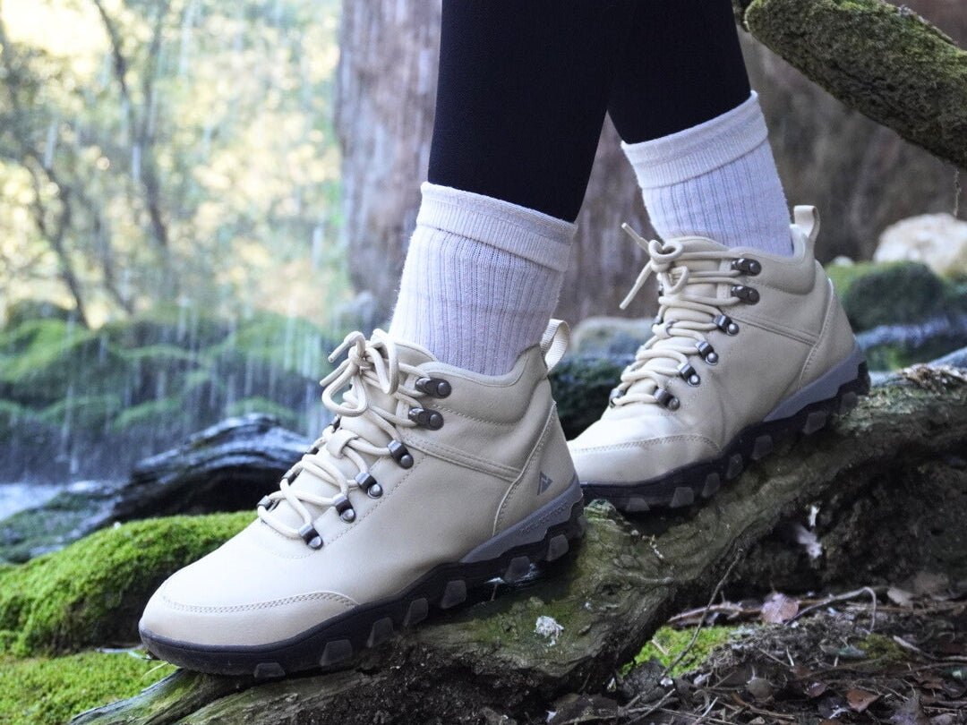 White hiking barefoot boots worn with black leggings and white socks, standing on a log in a forest setting.