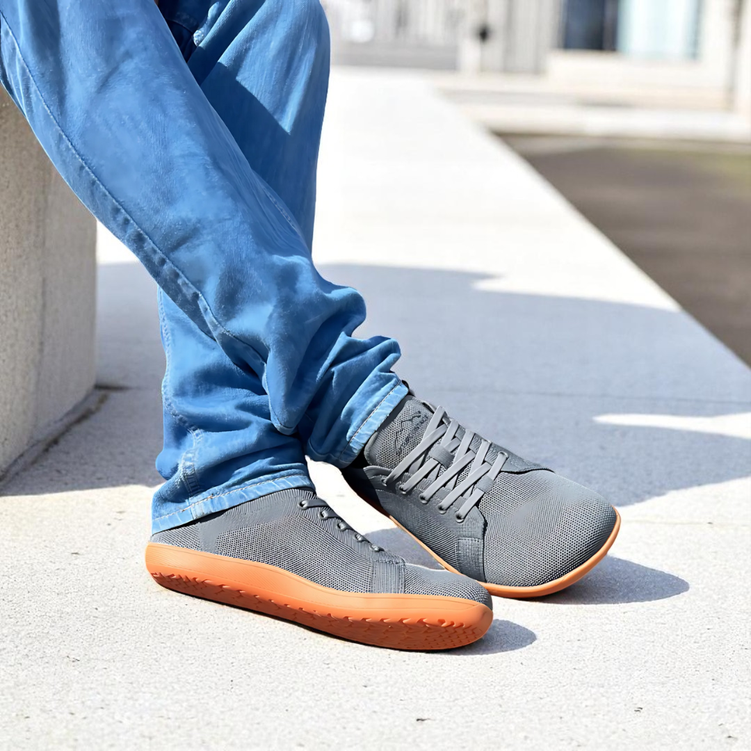 Gray sneakers with orange soles worn with blue jeans on a concrete surface.