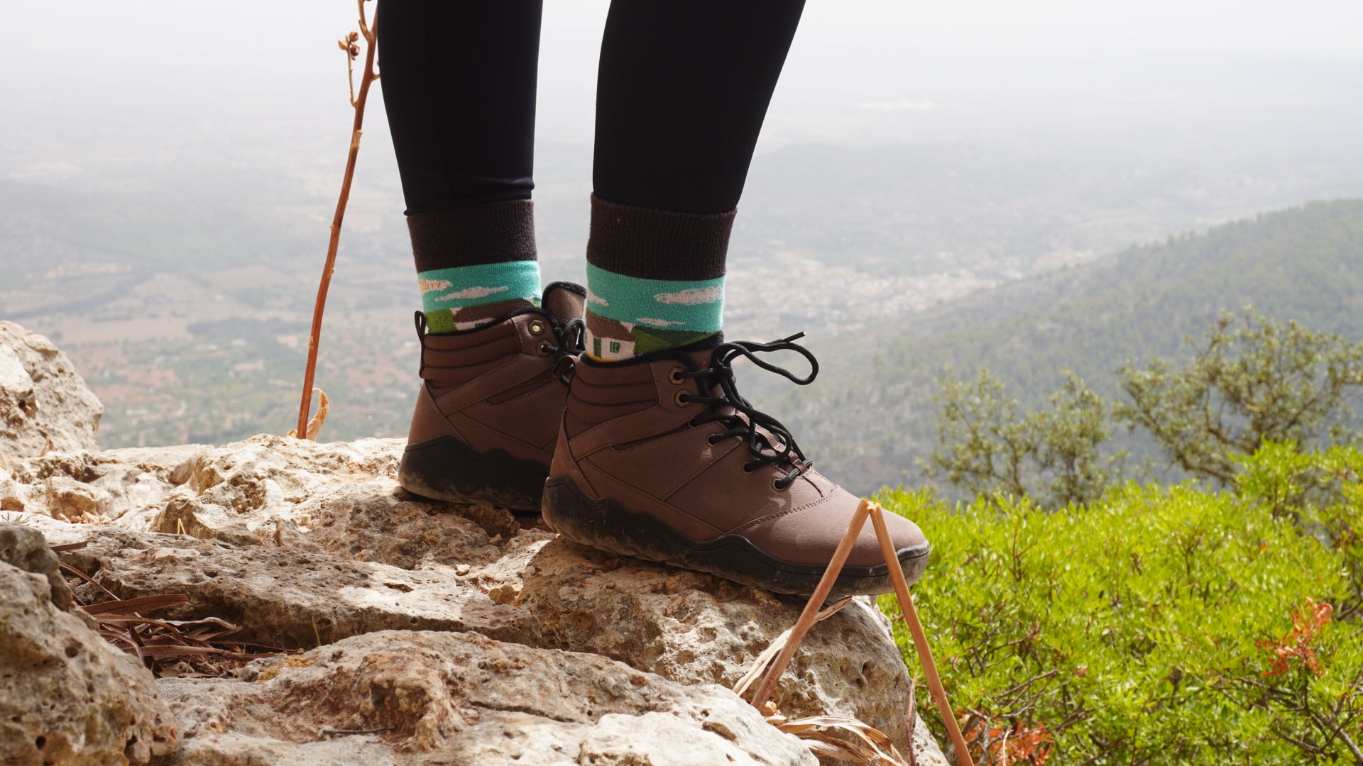 Person wearing brown hiking boots on a rocky outcrop with a scenic view of mountains and trees in the background.