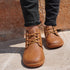 Brown leather shoes worn with black jeans against a stone wall.