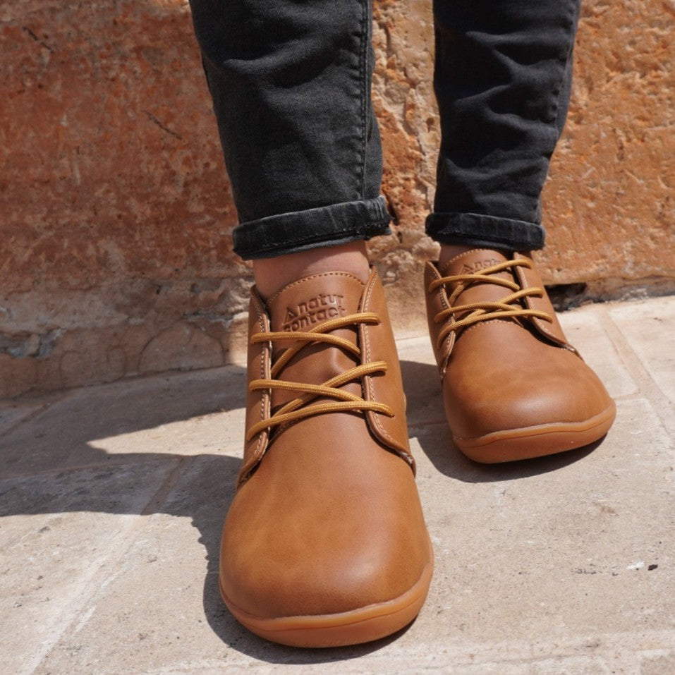 Brown leather shoes worn with black jeans against a stone wall.