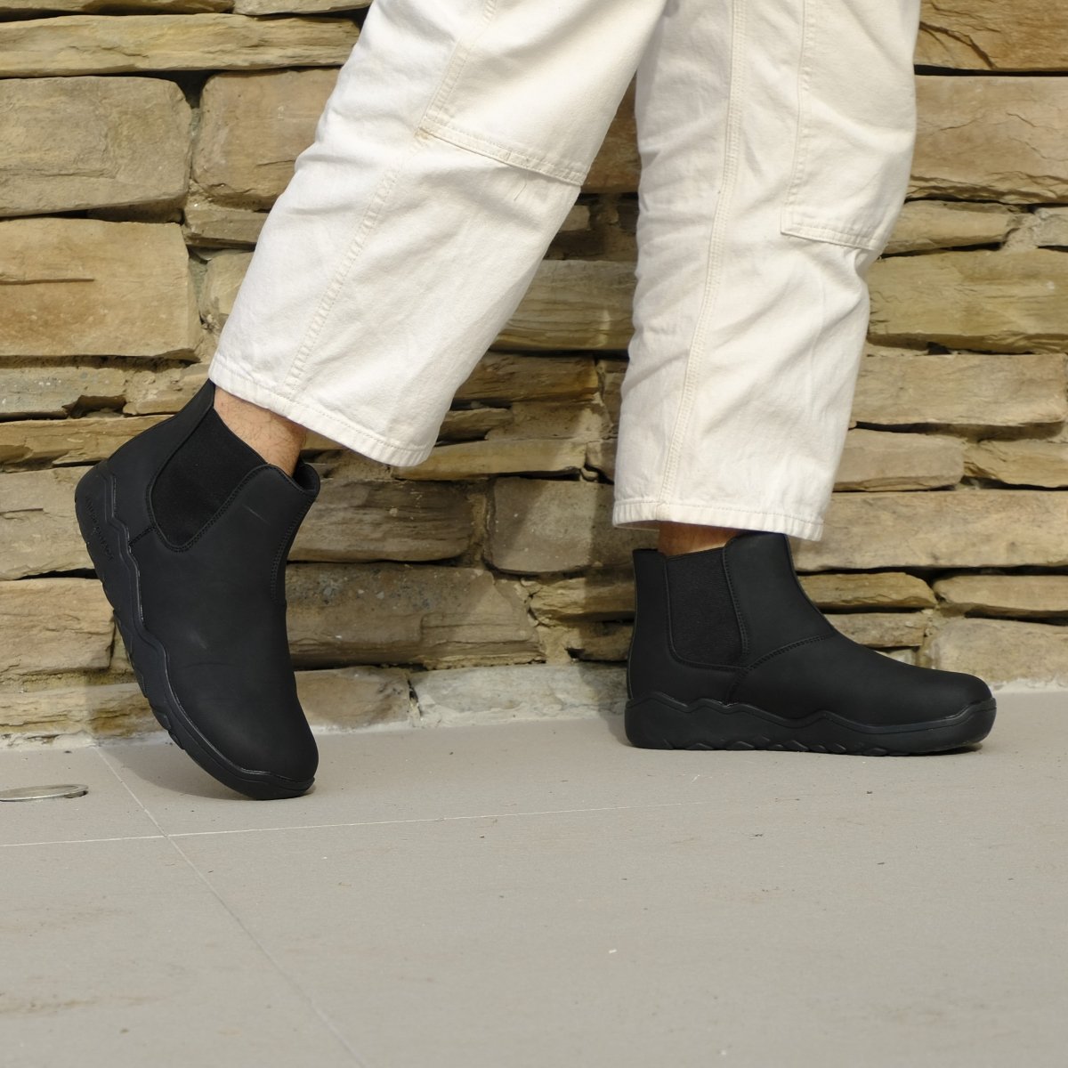 Person wearing black slip-on boots with white pants against a stone wall background