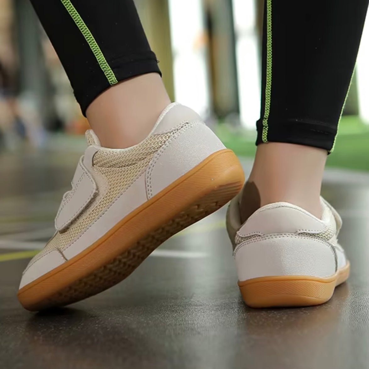beige barefoot sneakers for kids with brown soles worn by a person on a reflective surface.