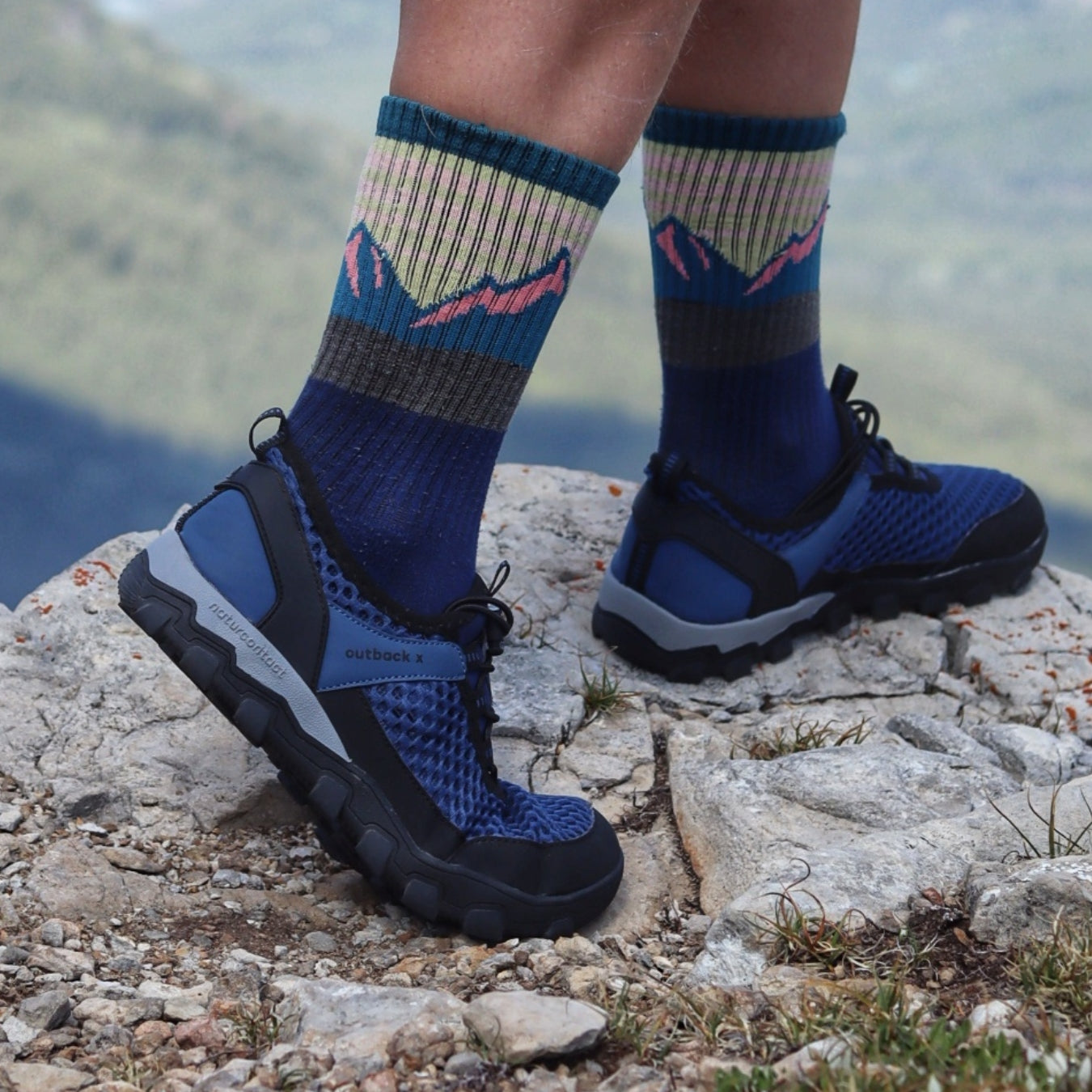 Person wearing blue hiking shoes and socks on a rocky surface with a mountainous background