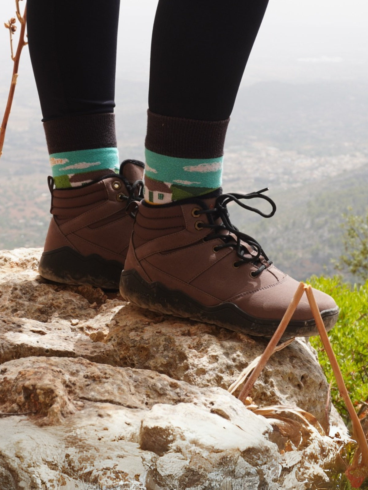 Person wearing brown boots and colorful socks standing on a rock with a mountainous background