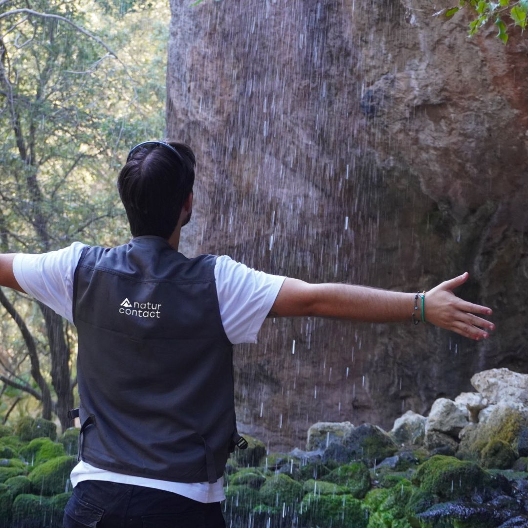 Person wearing a 'naturcontact' vest standing under a waterfall with arms outstretched.