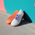 White barefoot shoes with orange soles and purple laces against a colorful wall.