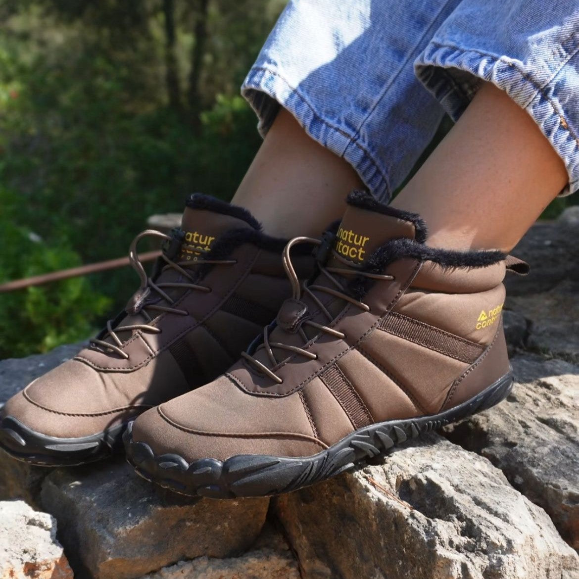 Brown hiking boots with 'natur tact' branding on a rocky surface with greenery in the background.