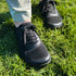Close-up of black mesh barefoot sneakers with laces, worn by a person standing on bright green grass.