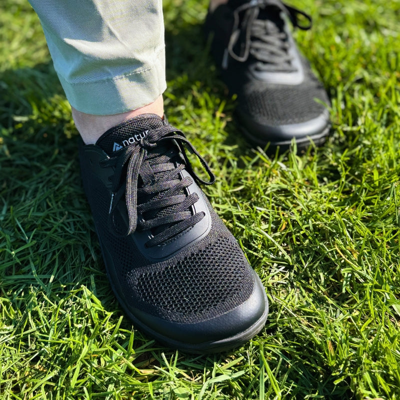 Close-up of black mesh barefoot sneakers with laces, worn by a person standing on bright green grass.
