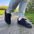 A person wearing grey rolled-up trousers and black mesh barefoot sneakers walking on a paved outdoor path during sunset.