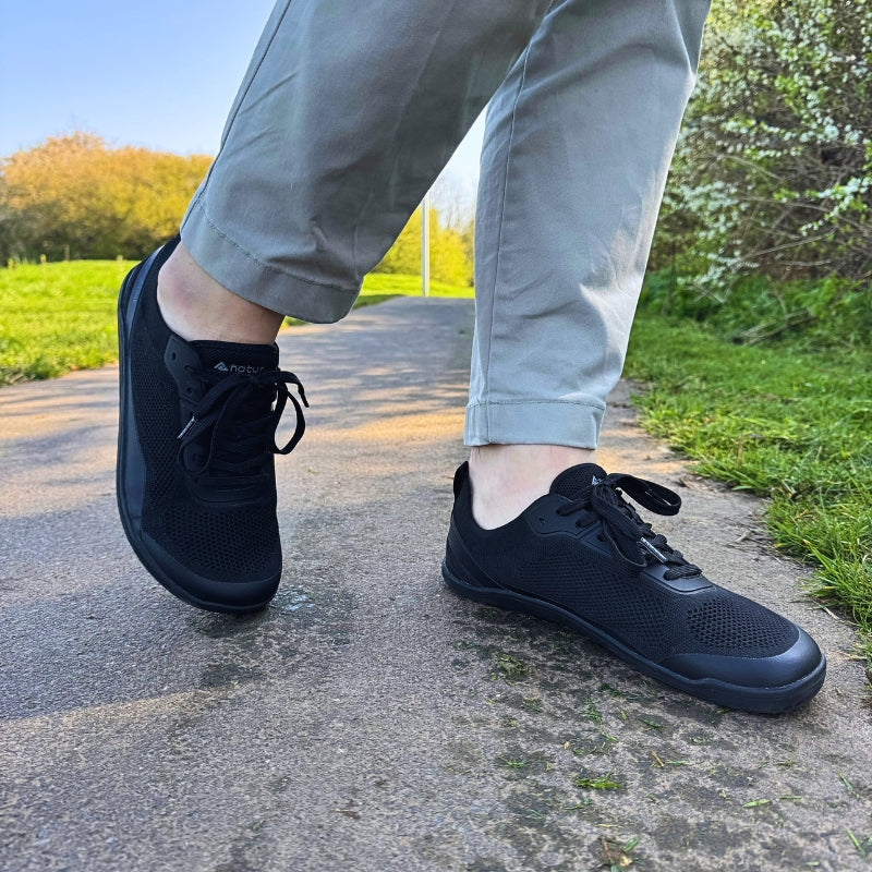 A person wearing grey rolled-up trousers and black mesh barefoot sneakers walking on a paved outdoor path during sunset.