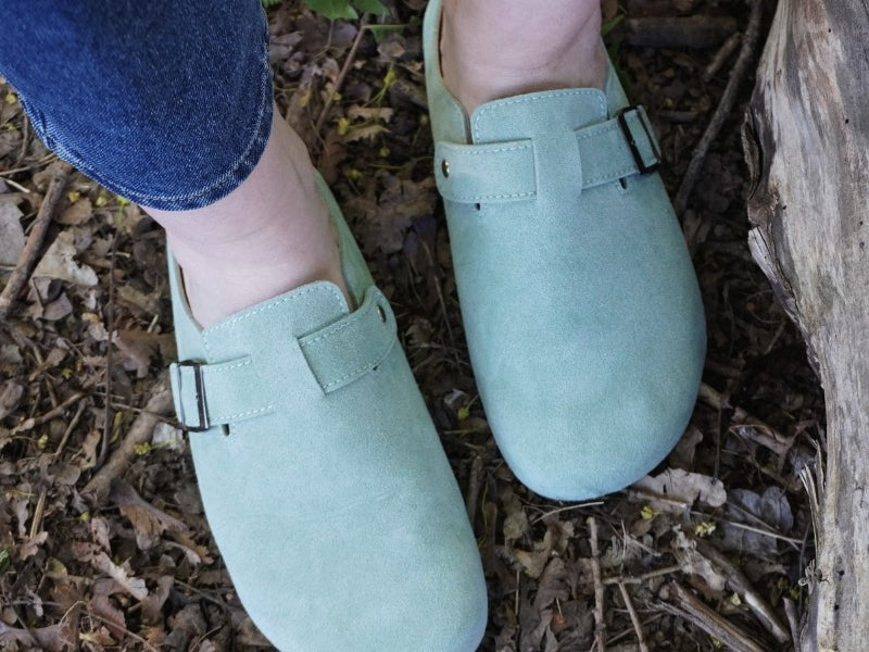 Light green clogs worn on a person's feet standing on dry leaves.