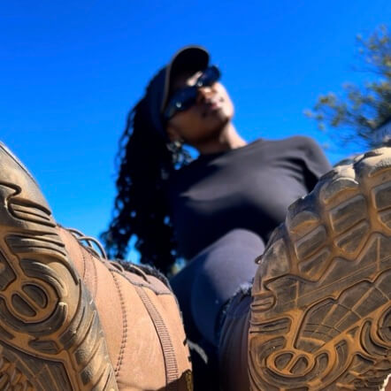 Person wearing a black outfit with a cap, standing between two large brown boots against a blue sky.