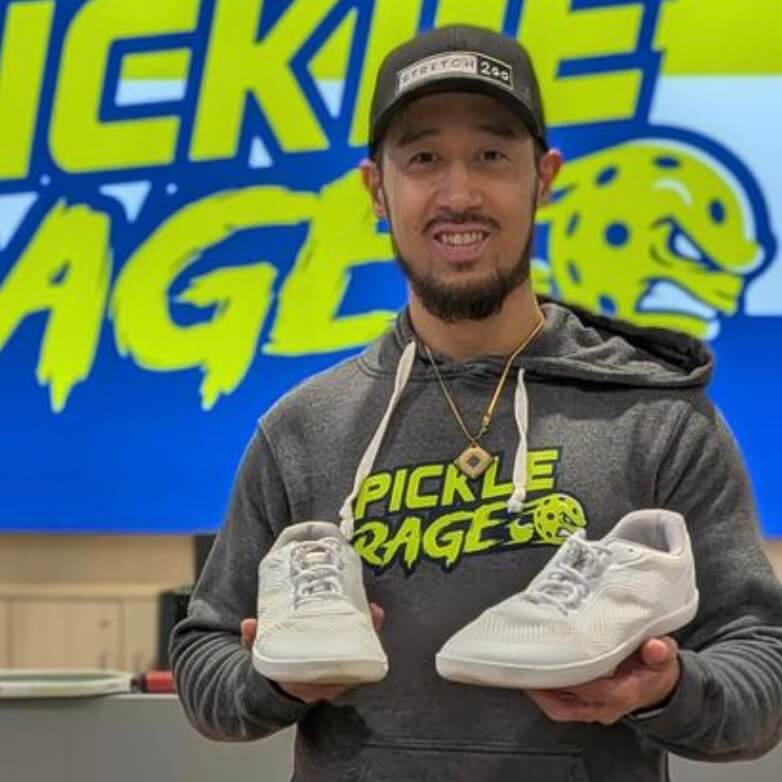 Person holding white barefoot trainers / sneakers in front of a Pickle Rage branded background