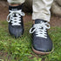 Front view of black leather barefoot sneakers with white laces, worn by a person standing on green grass.