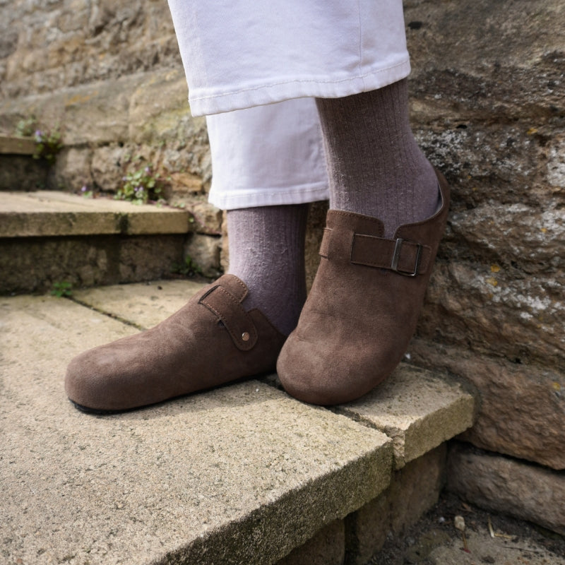 Close-up of brown suede clogs worn with thick grey socks on stone steps.
