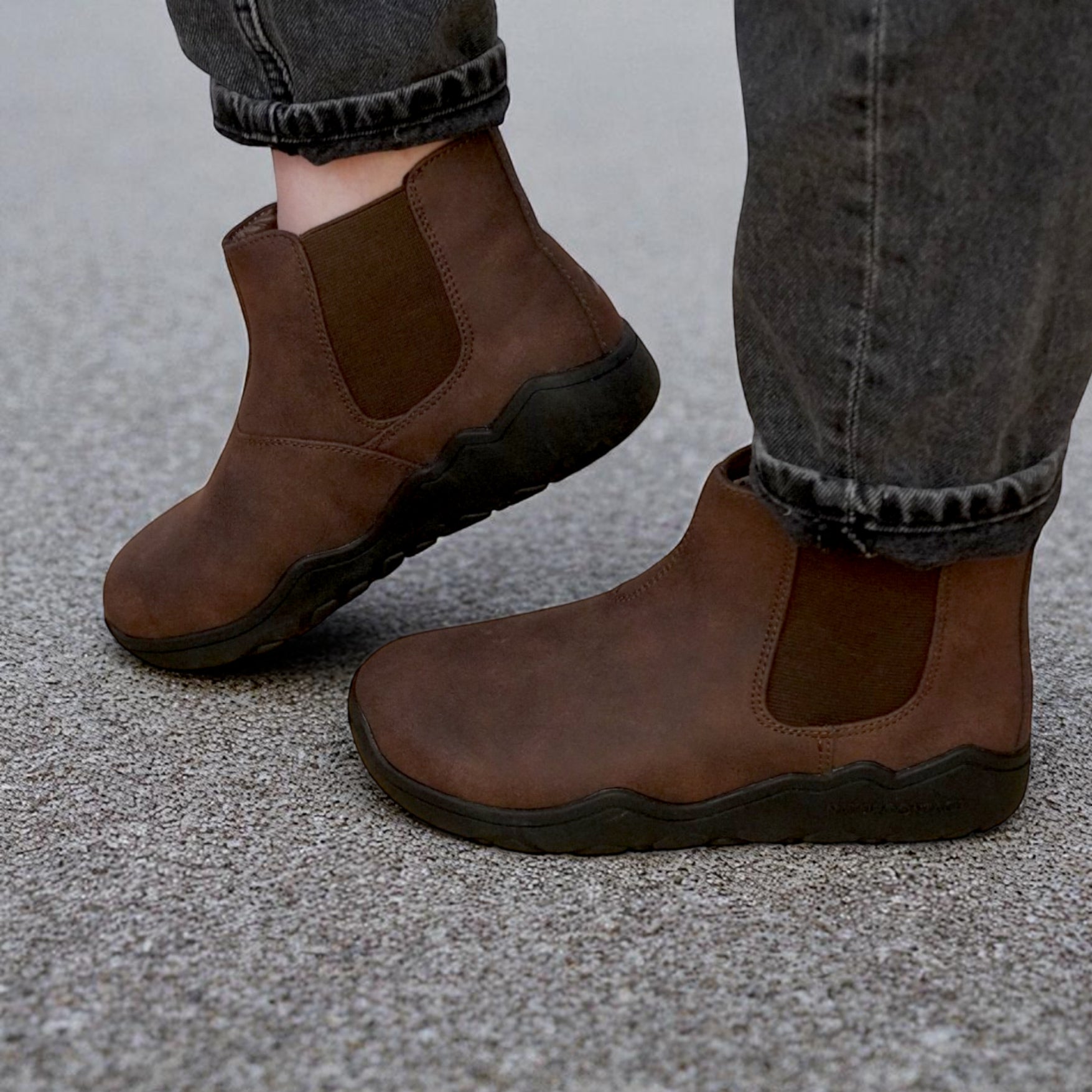 Brown ankle barefoot boots worn with gray jeans on a concrete surface