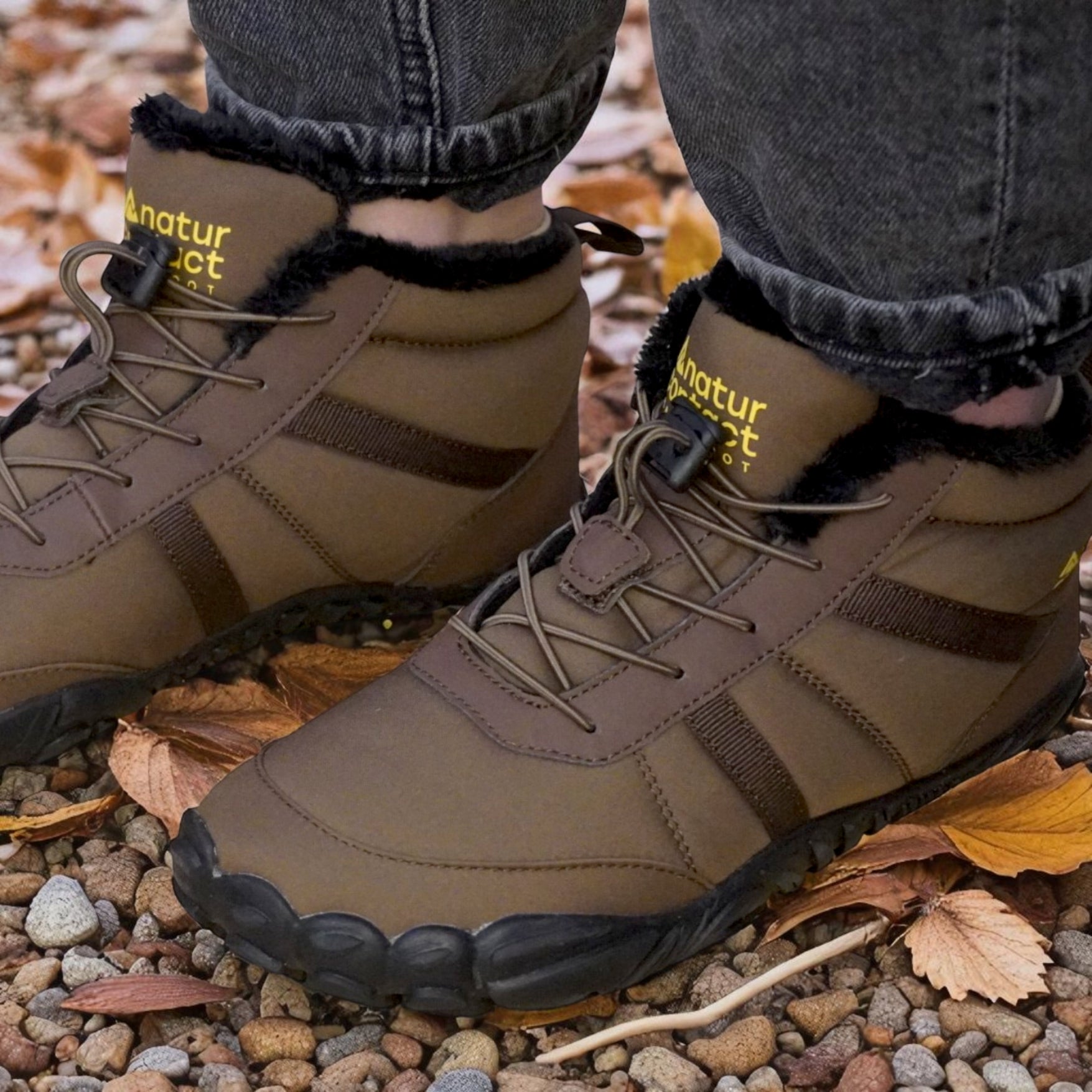 Brown boots with black soles worn on a rocky ground with autumn leaves.