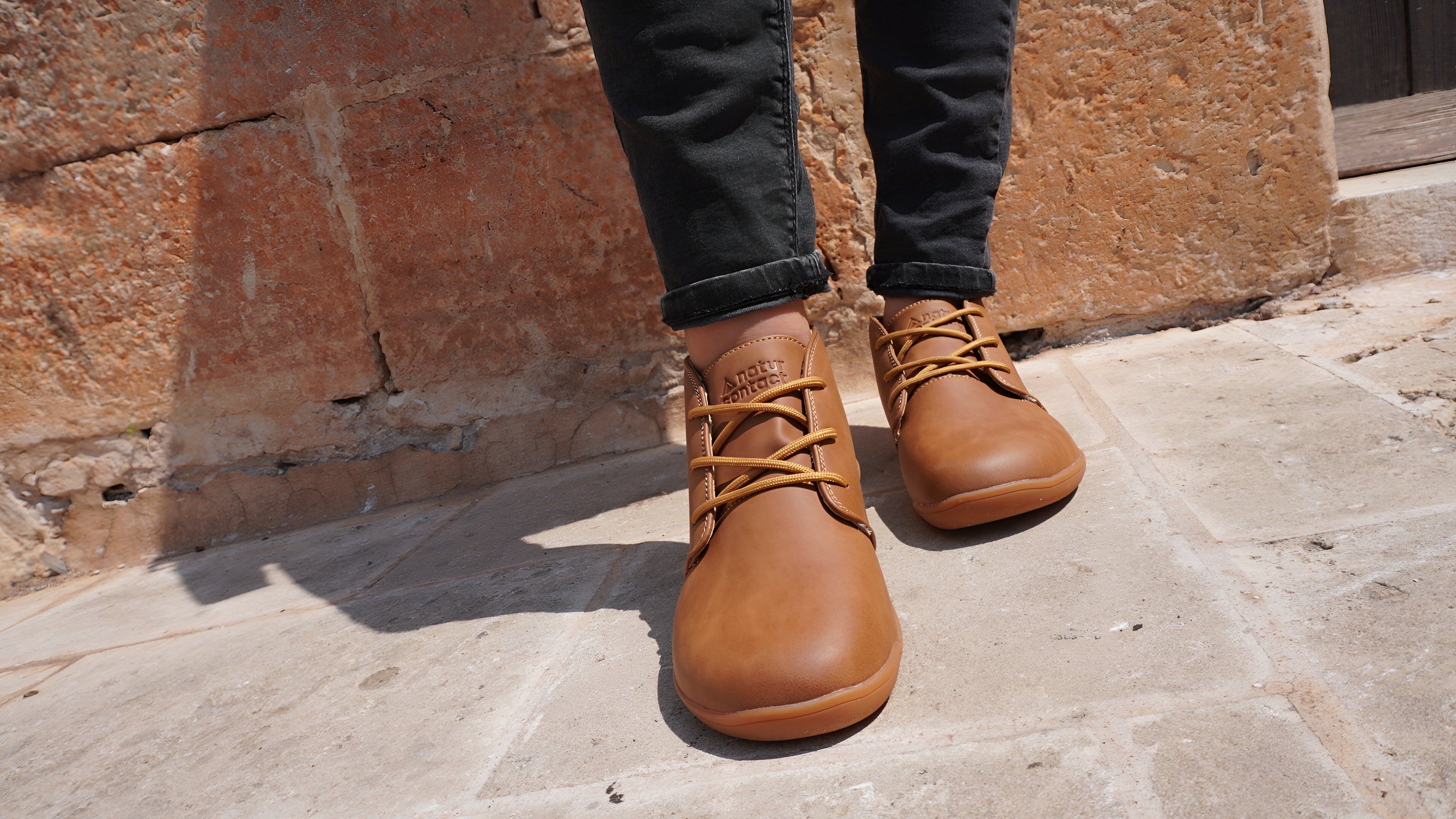 tobacco leather barefoot shoes worn with dark jeans against a stone wall.