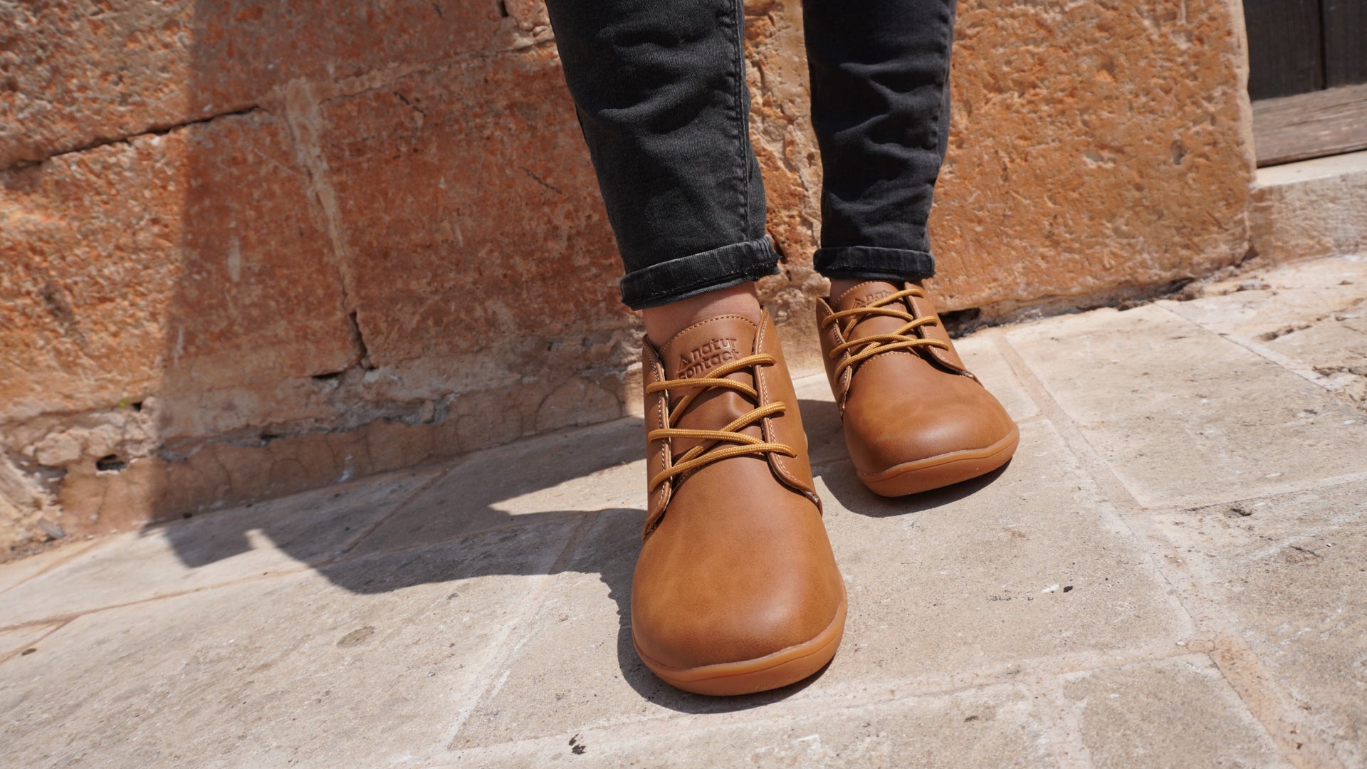 tobacco leather barefoot shoes worn with dark jeans against a stone wall.