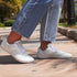White barefoot trainers sneakers worn with blue jeans on a sidewalk.