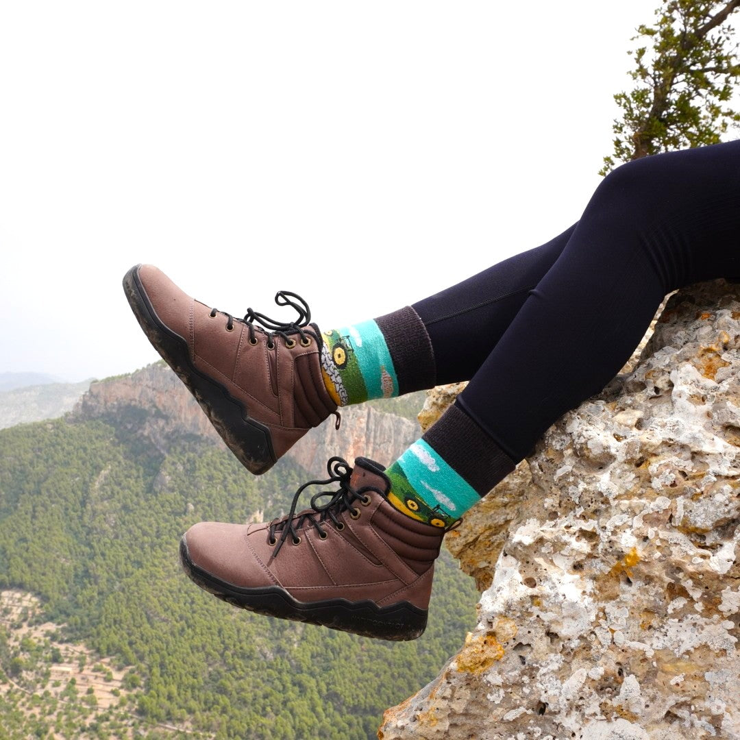 Brown hiking barefoot boots with black soles on a person sitting on a rock with a scenic background.