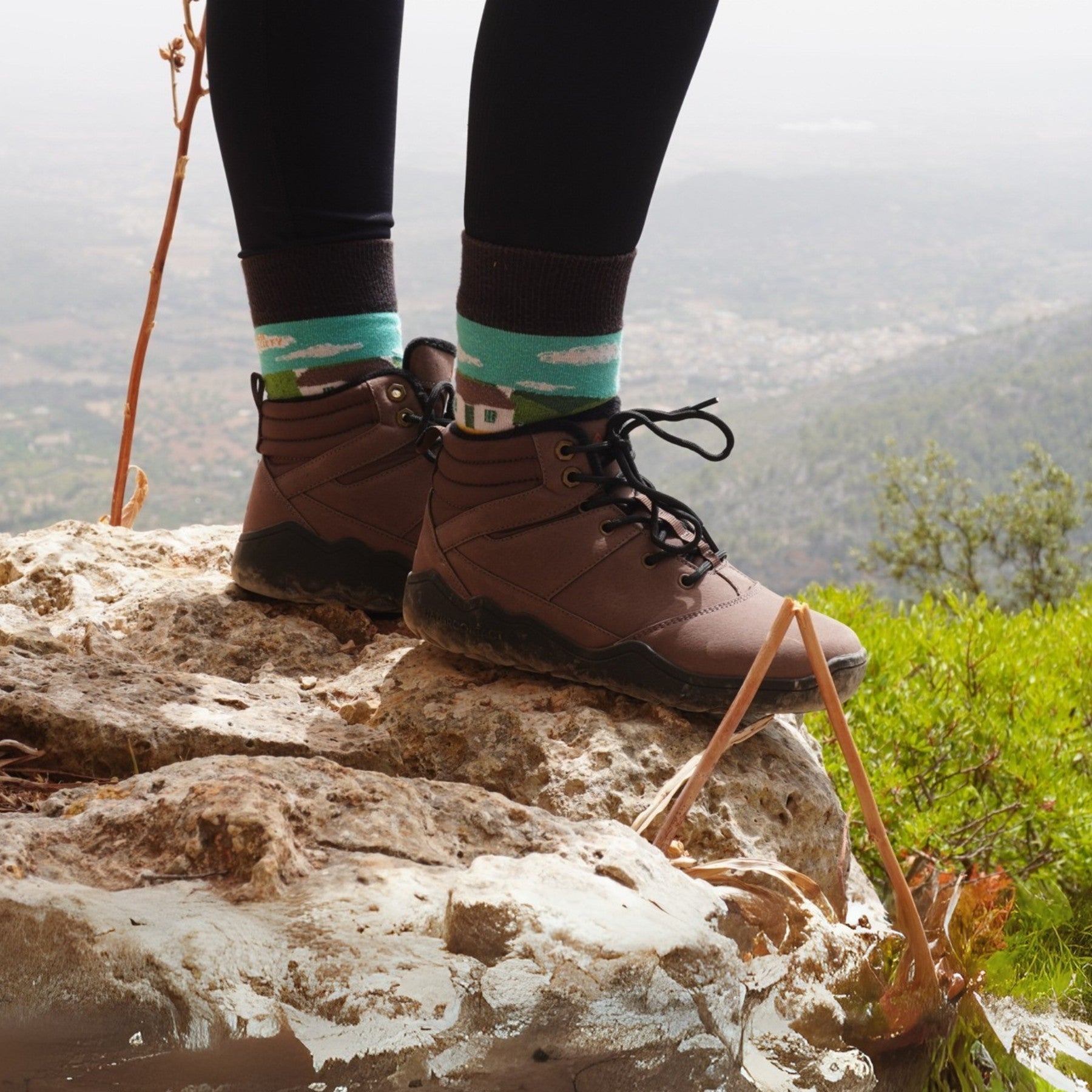Person wearing brown hiking barefoot boots and colorful socks standing on a rock with a scenic background