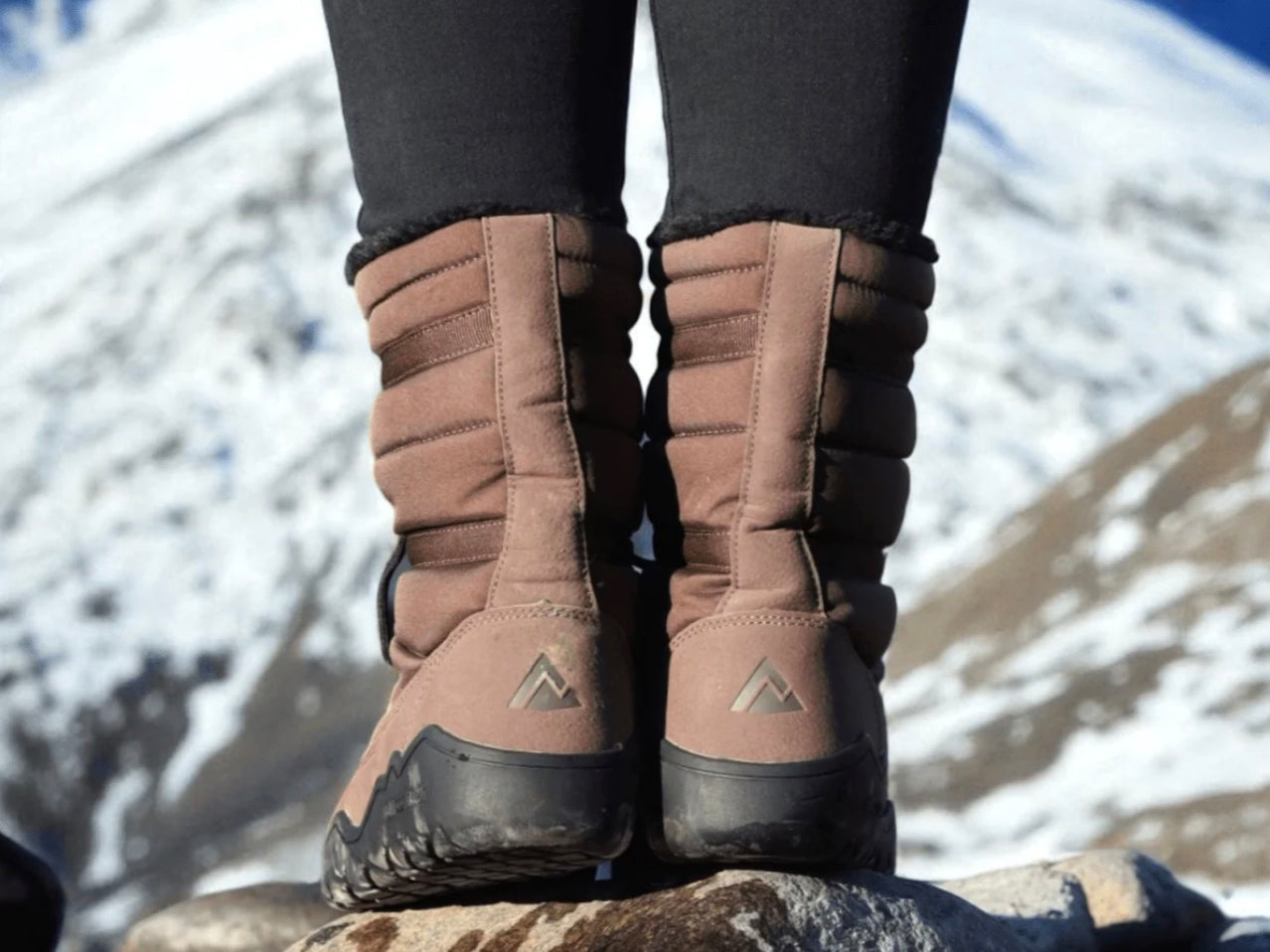 A person wearing mid-calf brown snowboard boots with black leggings, standing on rocks with a snowy mountain background.