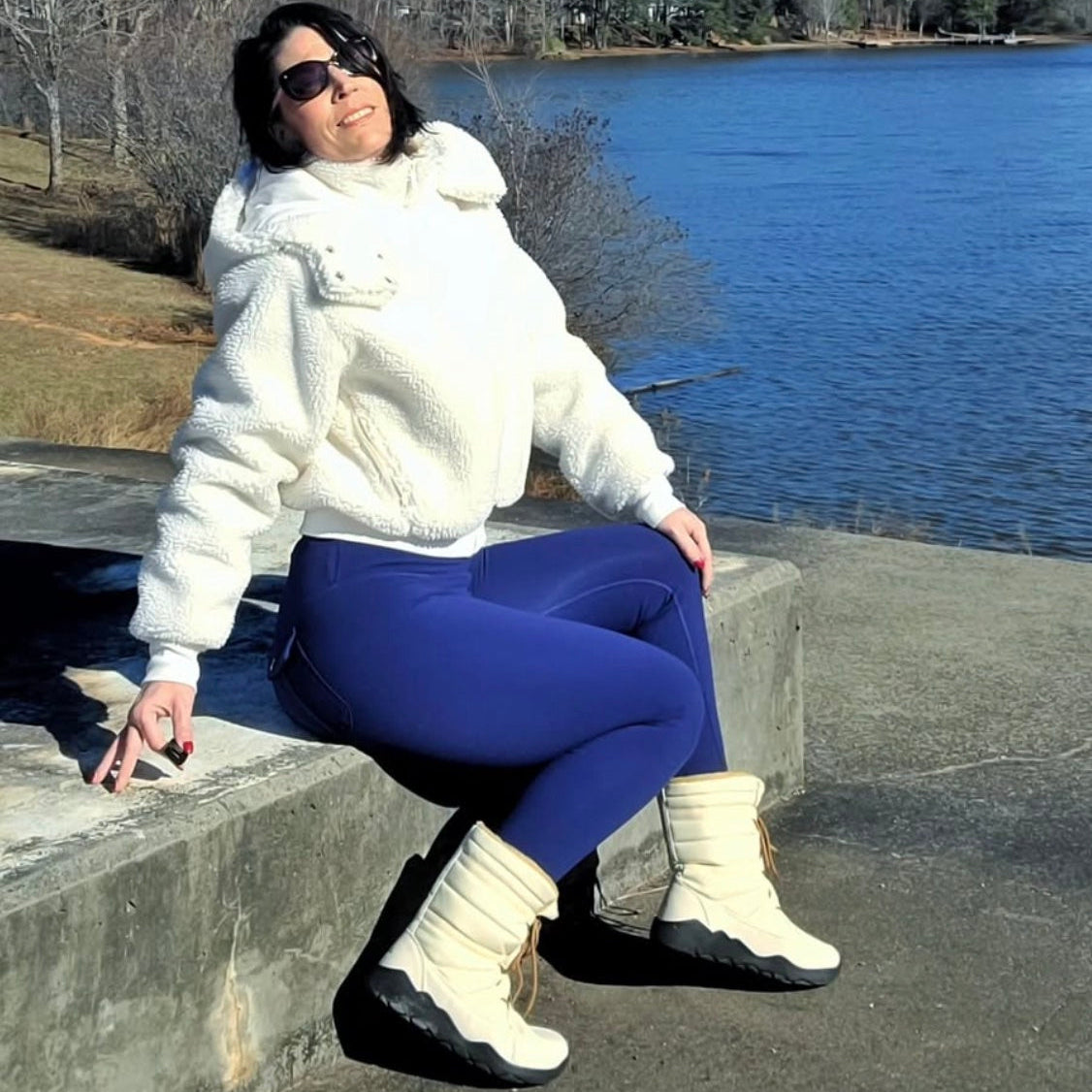 Person sitting by a lake wearing a white jacket, blue pants, and white boots.