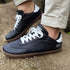 Side profile of black leather minimalist sneakers with gum soles and white laces, shown on a textured stone surface.