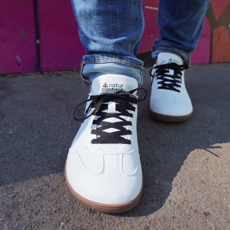Front close-up of white minimalist sneakers with black laces, worn with cuffed denim jeans against a purple wall background.