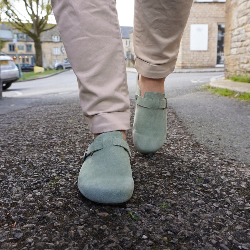 Person wearing mint green suede clogs and beige cuffed chinos on a cobblestone street.