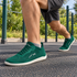 A person outdoors wearing dark green athletic sneakers with white soles and white zigzag logo details, shown in a running or lunge pose on a paved outdoor fitness area.