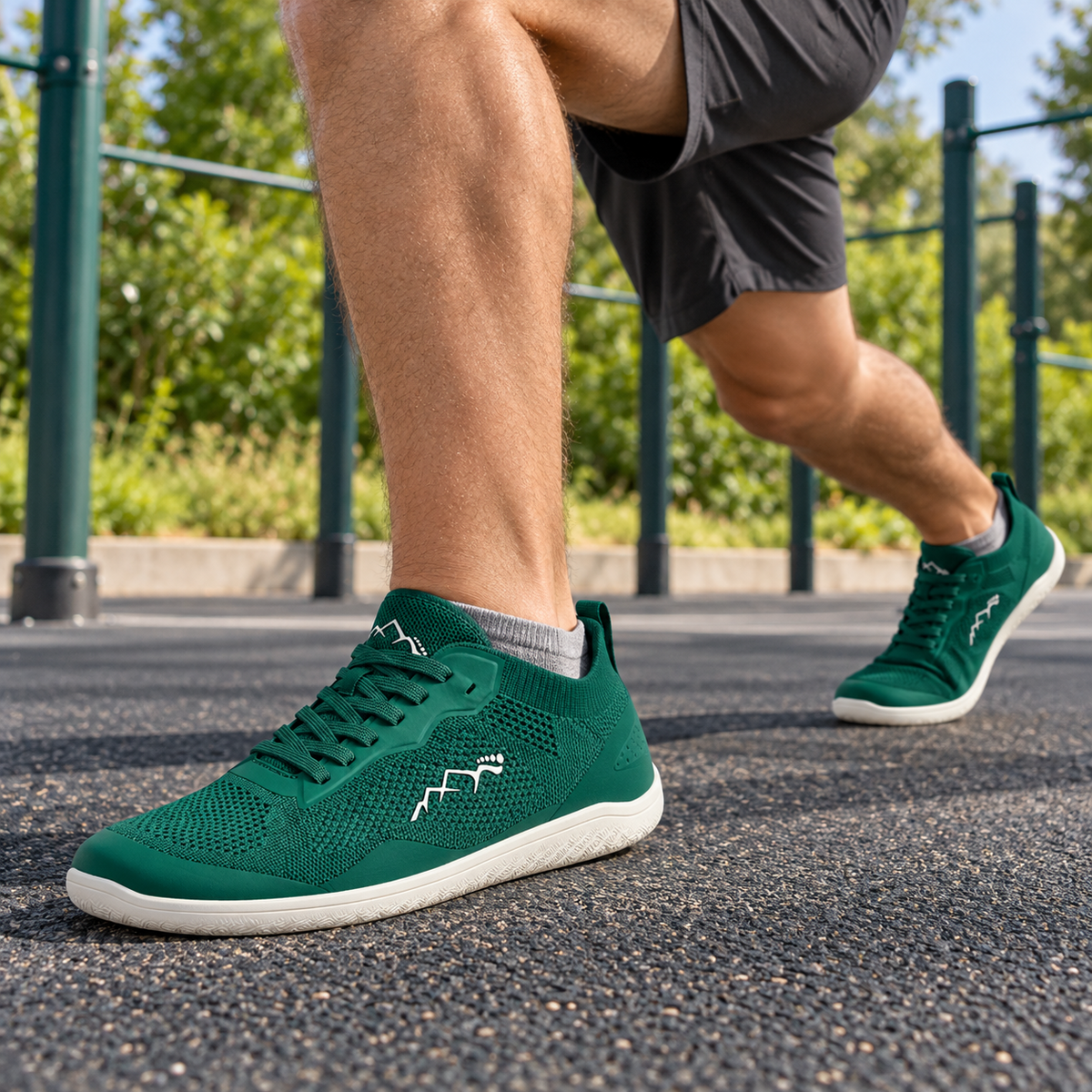 A person outdoors wearing dark green athletic sneakers with white soles and white zigzag logo details, shown in a running or lunge pose on a paved outdoor fitness area.