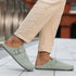 Light green barefoot suede clog loafers worn with beige pants on a blurred background