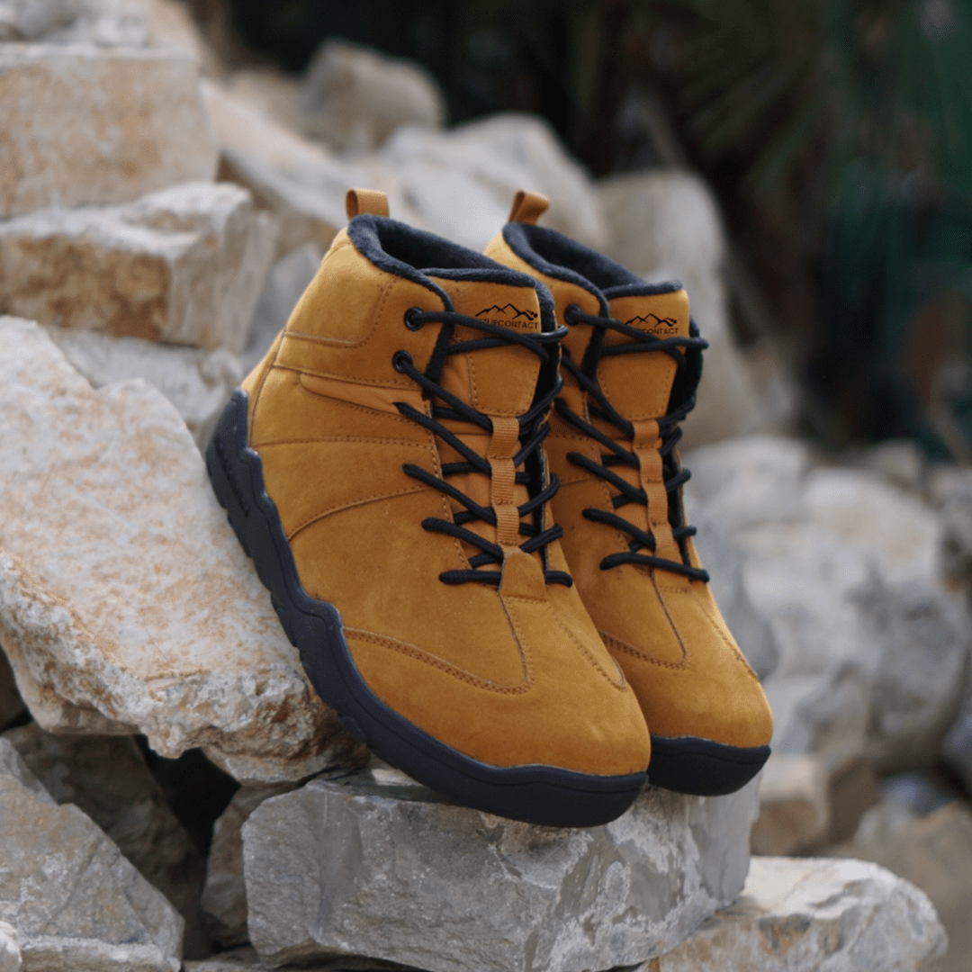 Pair of Naturcontact Intemperia winter barefoot boots in Gold color, with wide toe box, flexible sole and suede upper, placed on rocks in an outdoor setting.