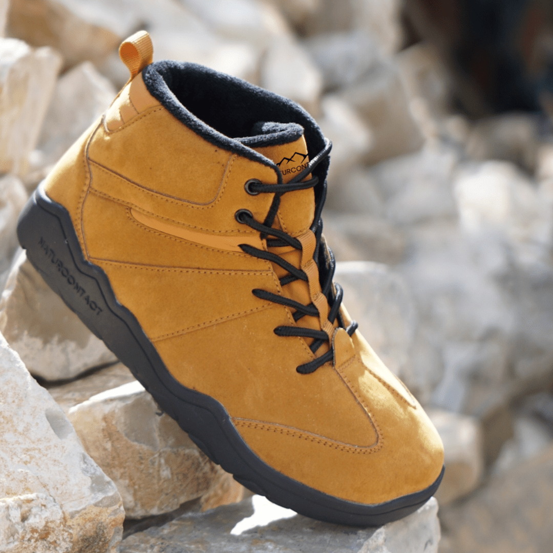 Naturcontact Intemperia winter barefoot boot in Gold color, waterproof minimalist design with wide toe box, shown on rocky terrain outdoors.