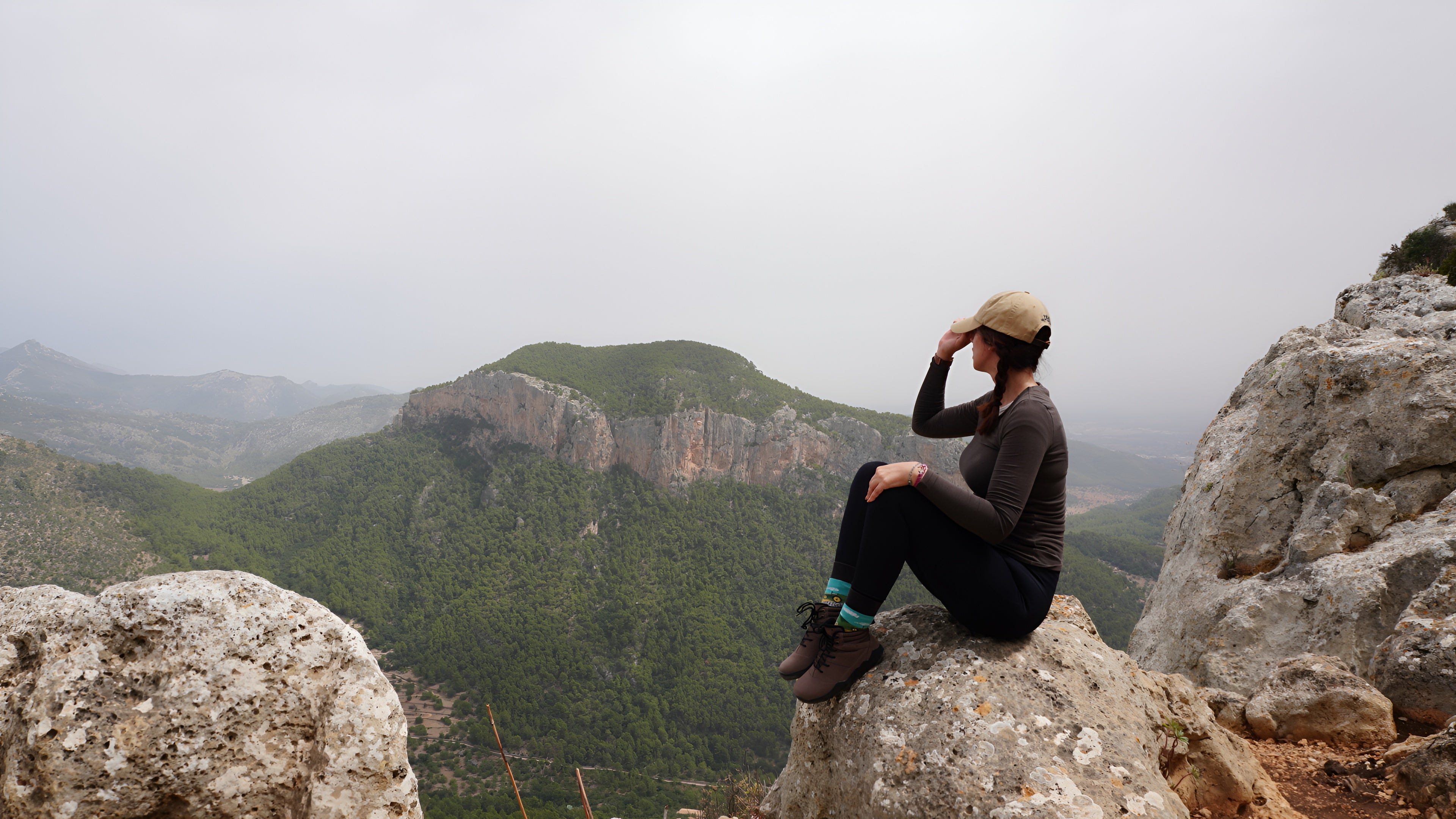 Person sitting on a rocky outcrop overlooking a mountainous landscape on a foggy day.