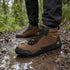 A person wearing brown and black waterproof hiking boots with black pants, standing on a muddy forest path with puddles. The boots feature a brown and black upper with yellow branding text.