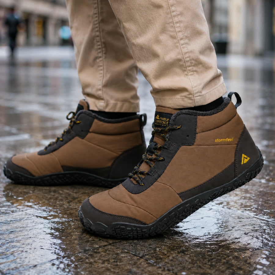 A person wearing tan and dark gray mid-top waterproof hiking boots with yellow branding, standing on a wet, reflective city street.