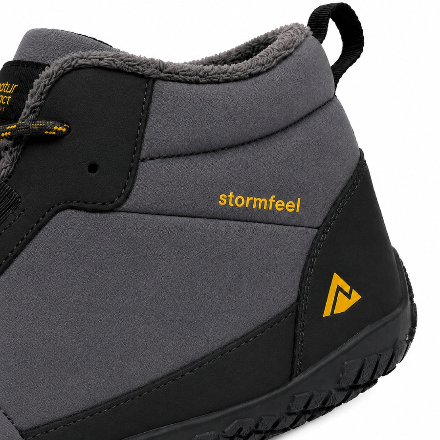 A close-up of a gray and black mid-height winter boot with yellow 'stormfeel' text and a yellow logo on the side, showing the heel, pull tab, and textured sole against a white background.
